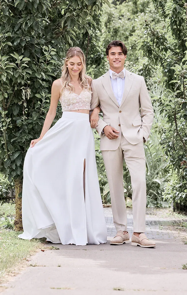 Sandfarbener Hochzeitsanzug mit hellbeiger Fliege, kombiniert mit leichter, sommerlicher Brautmode – stilvoller Boho-Look.