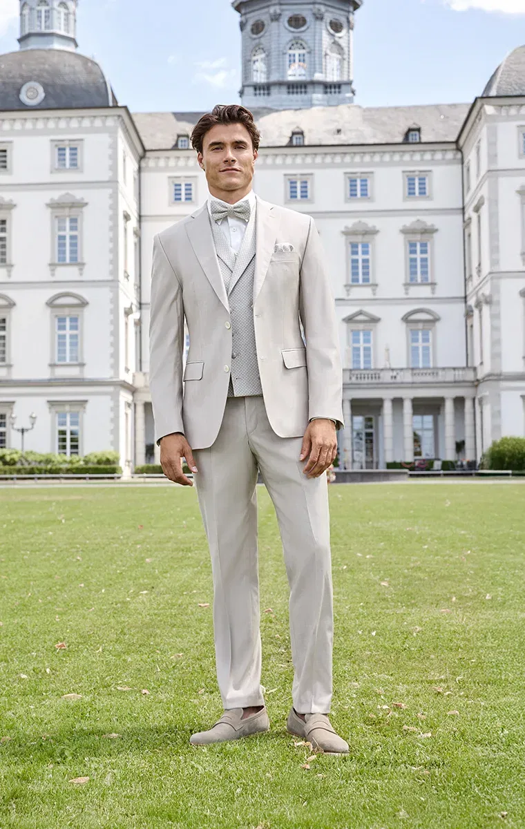 Bräutigam in cremefarbenem Anzug mit Weste und Fliege vor einem historischen Schloss – klassisch-eleganter Hochzeitslook.