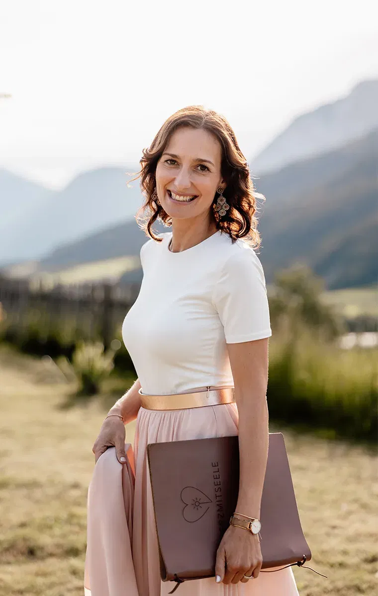 Frau im rosafarbenen Rock hält ein Traubuch mit Gravur „Herzmitseele“ – perfekte Kulisse in den Alpen.