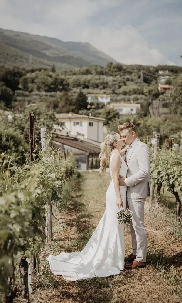 Ein Brautpaar küsst sich in einem sonnigen Weinberg mit Blick auf die Hügel und Häuser im Hintergrund.