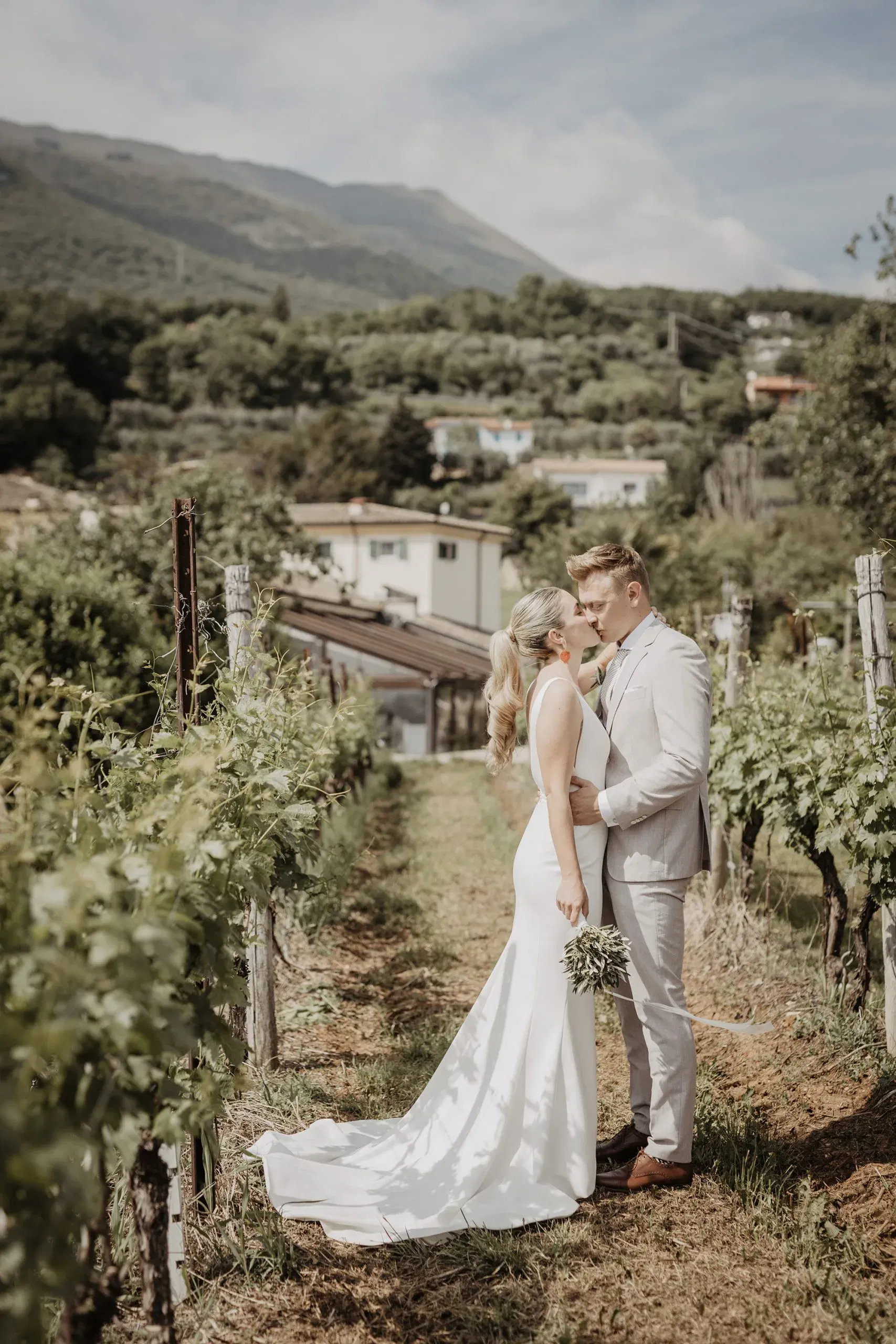 Ein Brautpaar küsst sich in einem sonnigen Weinberg mit Blick auf die Hügel und Häuser im Hintergrund.