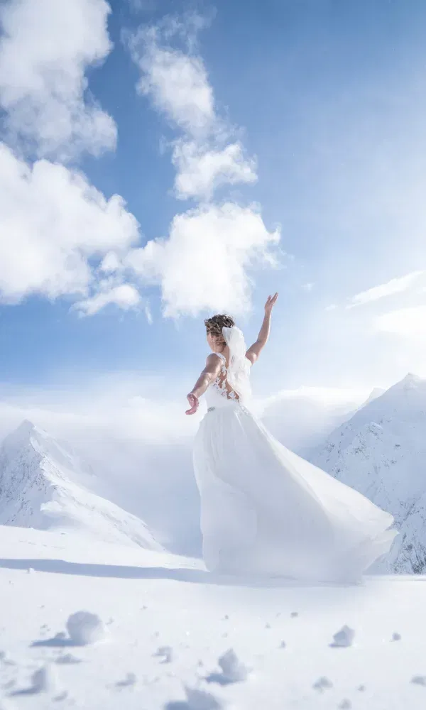 Anmutige Braut tanzt im Schnee vor atemberaubender Bergkulisse – ihr fließendes Kleid und der Schleier wehen im eisig-blauen Winterlicht.