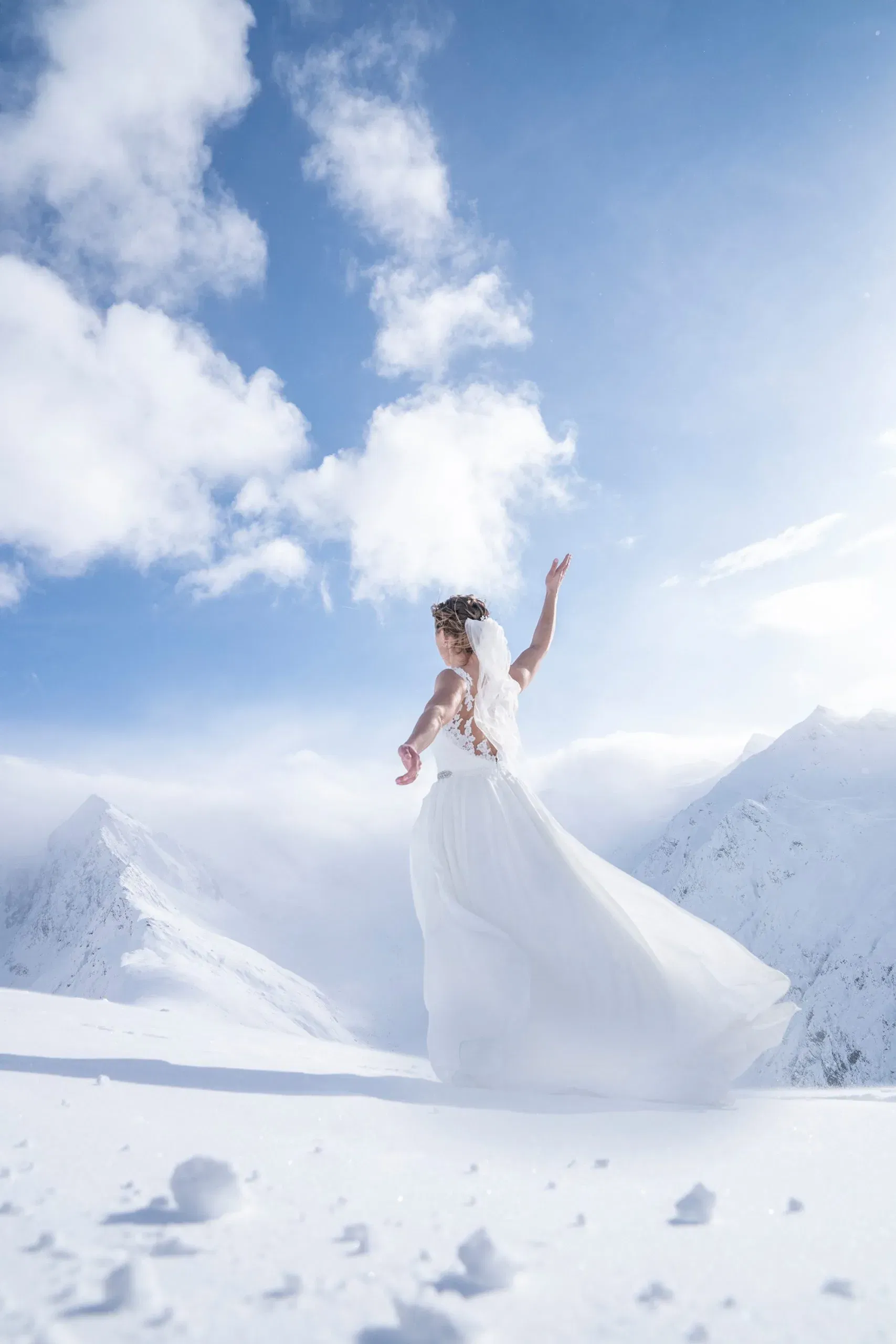 Anmutige Braut tanzt im Schnee vor atemberaubender Bergkulisse – ihr fließendes Kleid und der Schleier wehen im eisig-blauen Winterlicht.