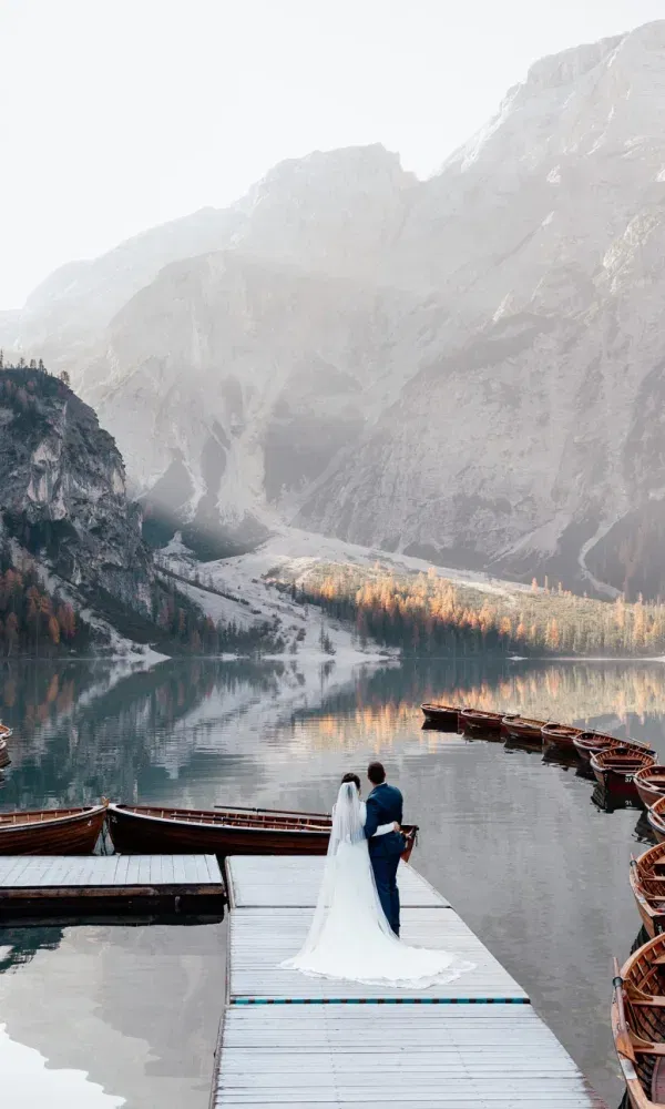Romantische Hochzeitsaufnahme eines Paares am stillen Bergsee mit Holzbooten und herbstlich gefärbtem Wald im Hintergrund.