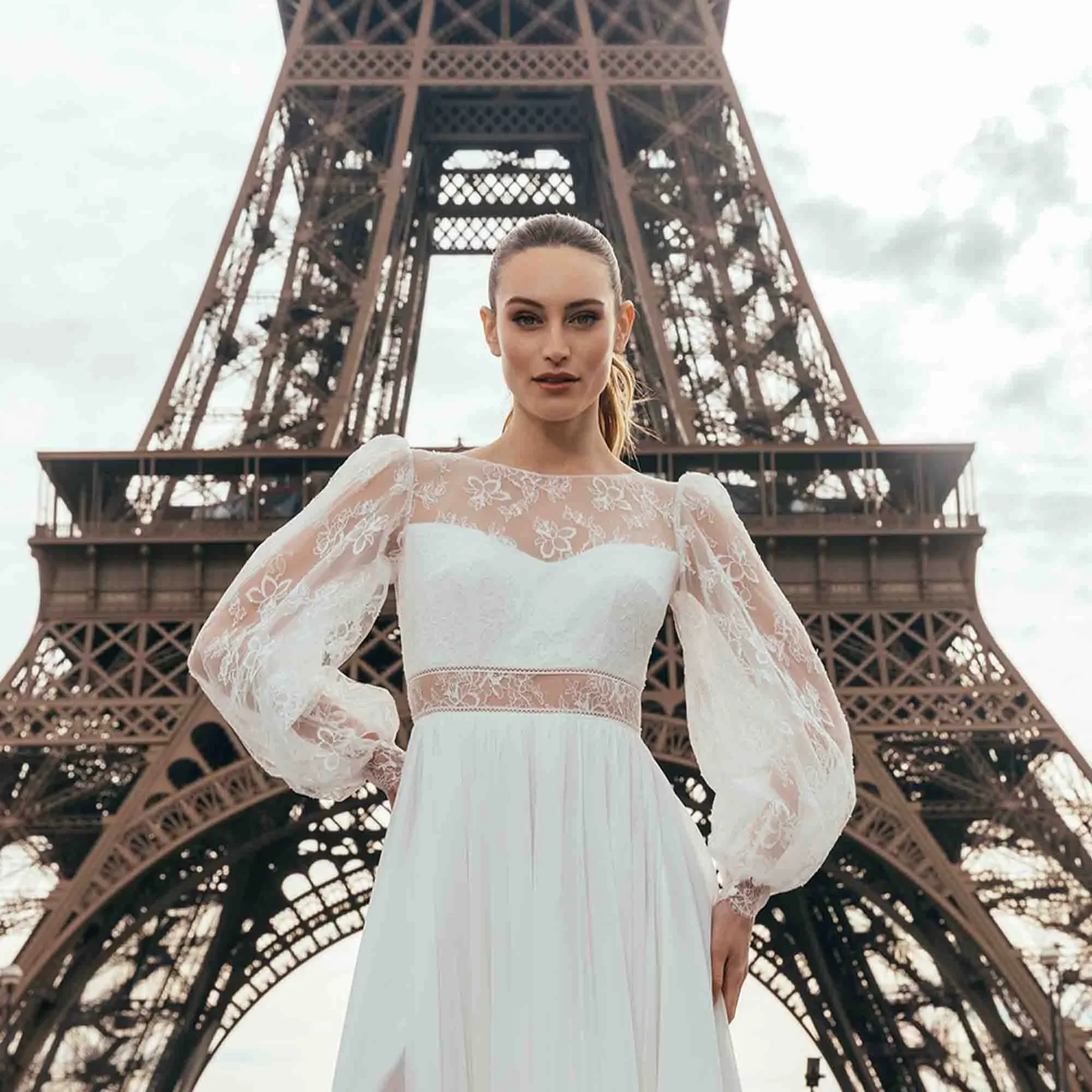 Braut in elegantem Spitzenkleid mit Puffärmeln posiert selbstbewusst vor dem Eiffelturm – Symbol einer modernen Power-Braut.
