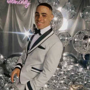 Junger Mann im hellgrauen Smoking mit schwarzen Akzenten und Fliege, lächelnd vor einer Kulisse aus glitzernden Spiegelballons und silbernem Hintergrund.