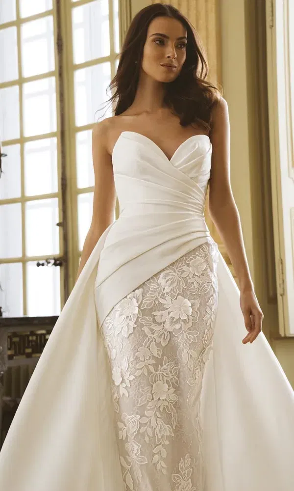 Elegantes trägerloses Brautkleid mit drapiertem Oberteil und floral besticktem Rock in hellem Interieur.