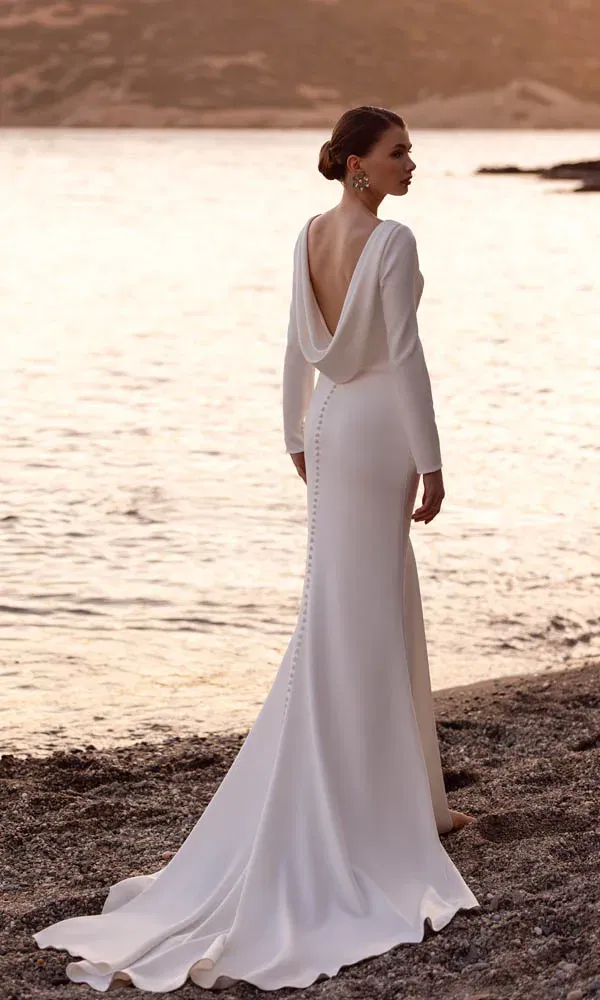 Elegantes Brautkleid mit tiefem Drapé-Rückenausschnitt und langer Schleppe, fotografiert am Strand bei Sonnenuntergang.