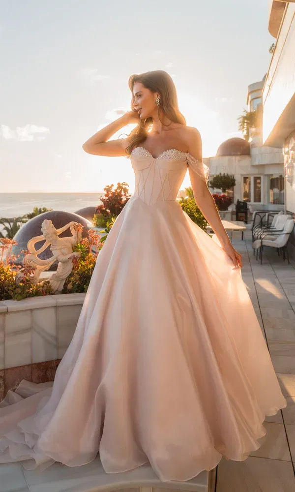 Romantisches Off-Shoulder-Ballkleid in zartem Blush mit Korsettoberteil, fotografiert im warmen Abendlicht am Meer.