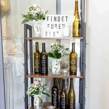 Kreativer Hochzeitssitzplan mit beschrifteten Weinflaschen und Blumenarrangements auf einem Regal.