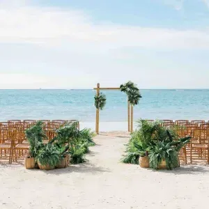 Trauungssetup am Strand mit Holzstühlen, grünem Palmschmuck und einem schlichten Traubogen vor dem Meer.