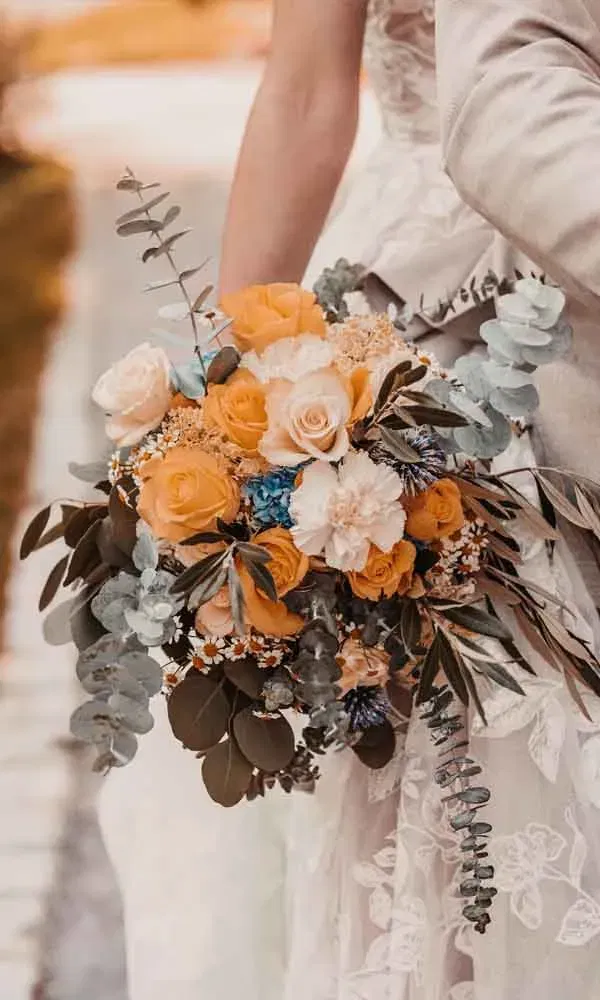 Romantischer Brautstrauß aus orangefarbenen Rosen, Cremeblüten und frischem Eukalyptus, getragen von einer Braut im Spitzenkleid.