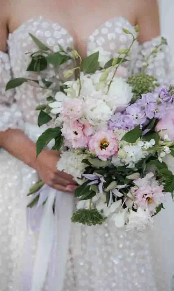 Zarter Brautstrauß in Pastellfarben aus Rosa, Weiß und Flieder, gehalten von einer Braut im perlenverzierten Kleid.