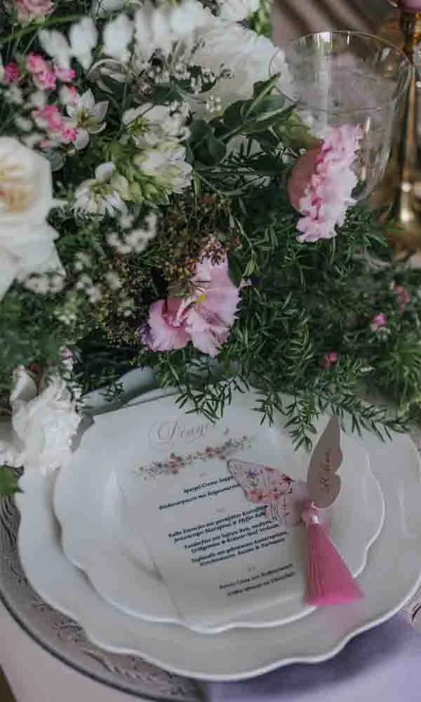 Festlich gedeckter Hochzeitstisch mit floraler Menükarte auf weißen Tellern, umgeben von üppigen rosa-weißen Blüten.