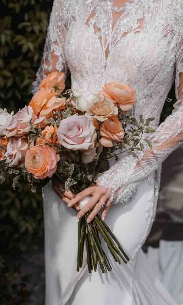Braut hält einen üppigen Brautstrauß in Pfirsich- und Rosétönen, dazu ein detailreiches Spitzenkleid.