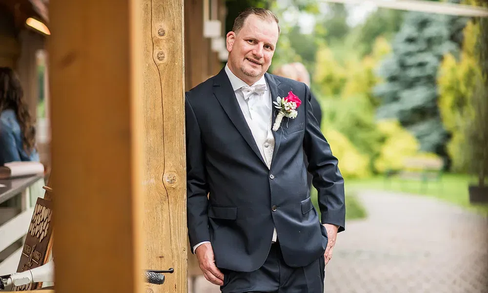 Der Bräutigam lehnt entspannt an einem Holzpfosten und lächelt in die Kamera, umgeben von einer grünen Gartenkulisse.