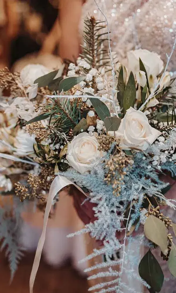 Winterlicher Brautstrauß mit weißen Rosen, Eukalyptus, Tannenzweigen und zarten Trockenblumen, gehalten von einer Braut im Glitzerkleid.