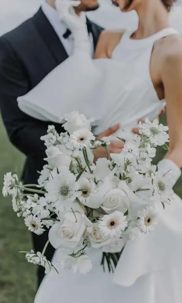 Elegant gekleidete Braut hält einen modernen weißen Brautstrauß aus Rosen und Gerbera, während sie zärtlich vom Bräutigam umarmt wird.
