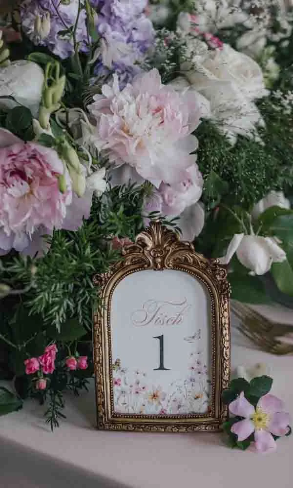 Eleganter Rahmen mit Tischnummer, eingebettet in ein reiches Blumenarrangement aus Pfingstrosen, Rosen und Grün.