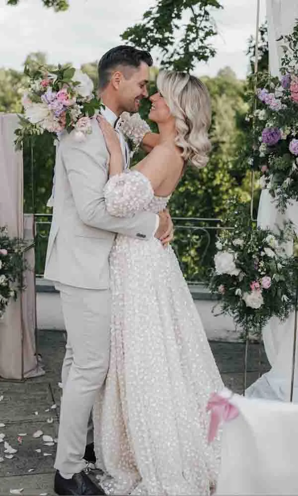 Ein Brautpaar umarmt sich liebevoll vor einer blumengeschmückten Outdoor-Kulisse; die Braut trägt ein helles, strukturiertes Kleid, der Bräutigam einen hellen Anzug.