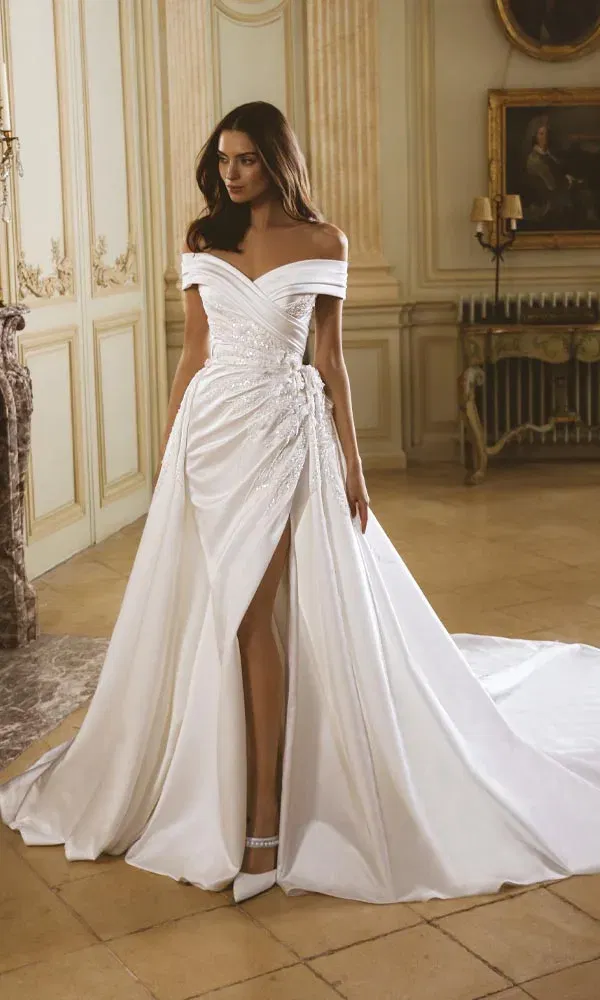 Opulentes Off-Shoulder-Brautkleid mit drapiertem Oberteil, Perlendetails und hohem Schlitz in eleganter Schlosskulisse.