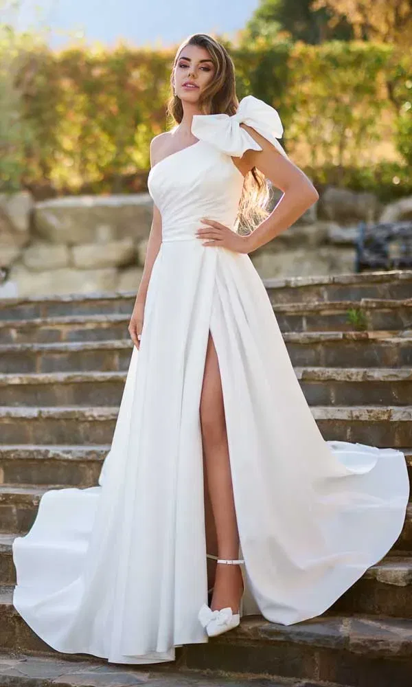 Braut in einem One-Shoulder-Kleid mit großer Schleife und hohem Schlitz auf einer Steintreppe.