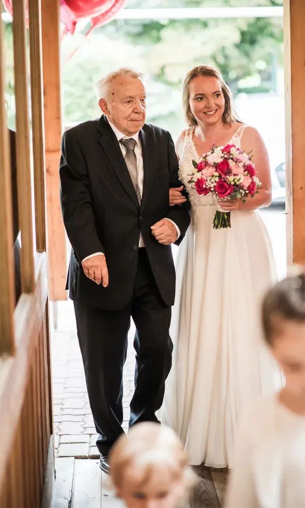 Eine Braut schreitet Arm in Arm mit einem älteren Mann durch den Eingang, während sie ihren farbenfrohen Blumenstrauß trägt.