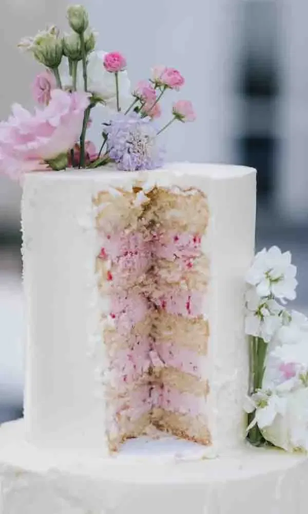 Weiße Hochzeitstorte mit floralem Topping, bei der ein hohes Stück herausgeschnitten wurde und die rosa Cremefüllung sichtbar ist.
