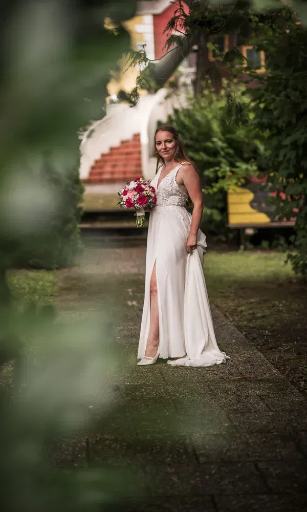 Die Braut steht mit ihrem Blumenstrauß auf einem Gartenweg und blickt lächelnd durch eine natürliche grüne Blätterumrahmung in die Kamera.