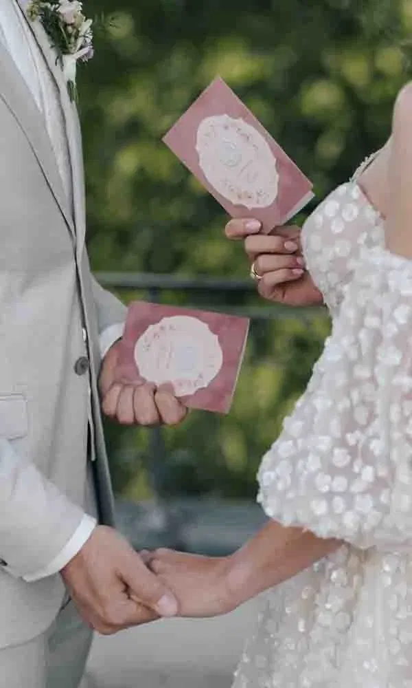 Braut und Bräutigam halten während der Trauung ihre persönlichen Eheversprechen in rosafarbenen Heftchen und fassen sich an den Händen.