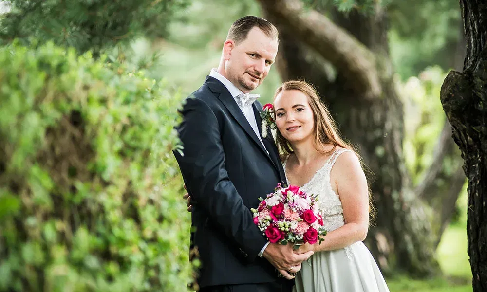 Braut und Bräutigam stehen eng umarmt im Garten, während sie gemeinsam den bunt gemischten Brautstrauß halten.