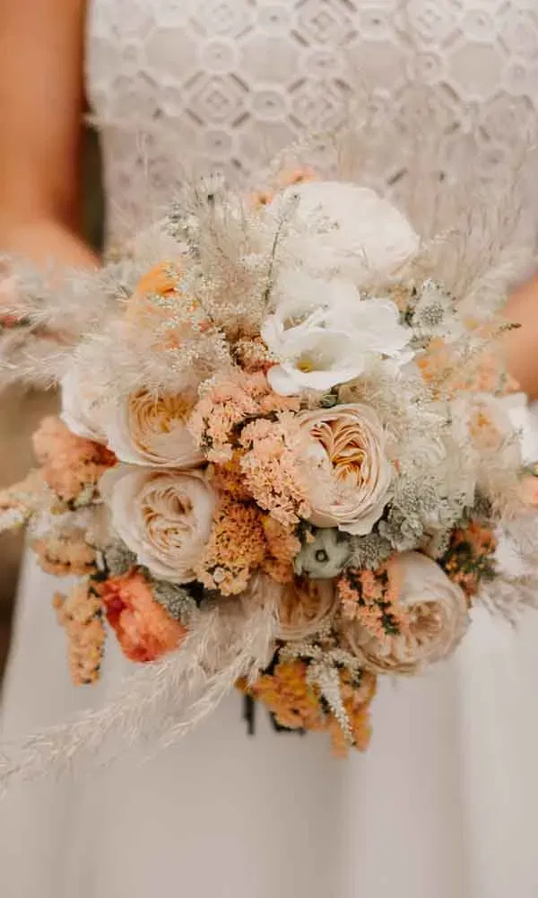 Braut hält einen romantischen Brautstrauß in Pfirsich- und Cremetönen mit Rosen, Trockenblumen und Pampasgras.