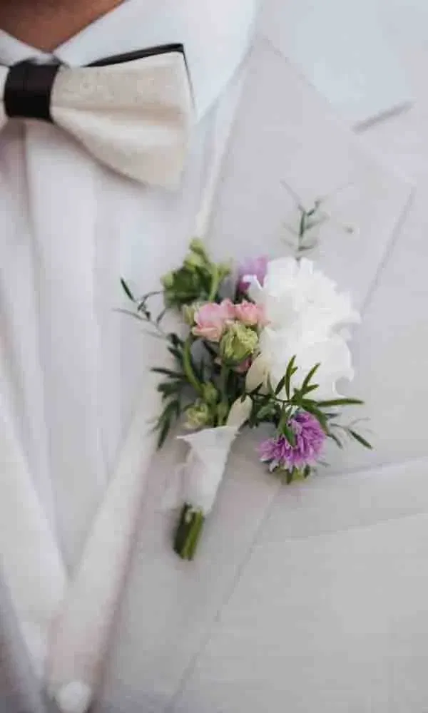 Nahaufnahme eines hellen Anzugs mit cremefarbener Fliege und einer zarten Ansteckblume aus weißen, rosa und violetten Blüten.