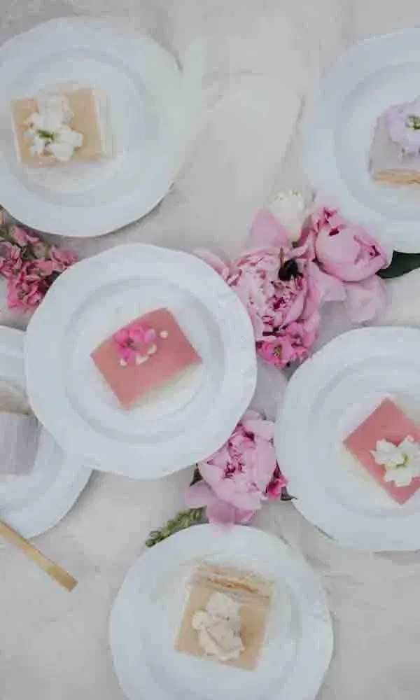 Von oben fotografierte Dessertteller mit kleinen Kuchenstücken in Rosa- und Cremetönen, umrahmt von pinken Pfingstrosen.