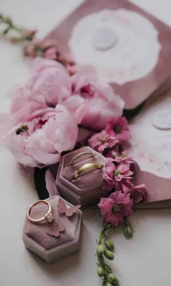 Detailaufnahme von goldenen Trauringen und einem Verlobungsring in roséfarbenen Ringboxen, umgeben von pinken Pfingstrosen und romantischer Papeterie.