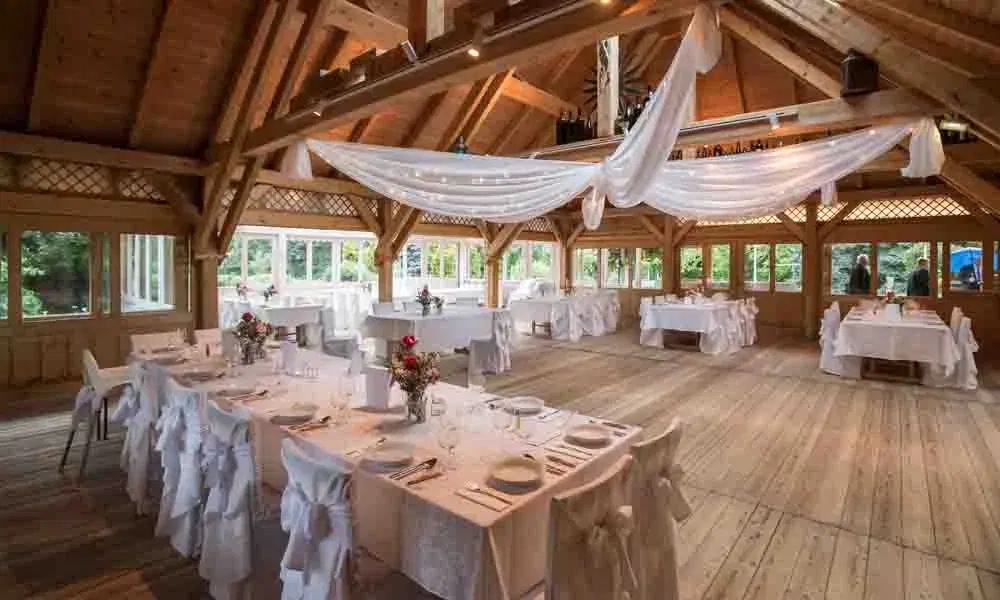 Festlich dekorierter Hochzeitssaal mit weiß eingedeckten Tischen und eleganten Stoffbahnen, die unter dem Holzdach schweben.