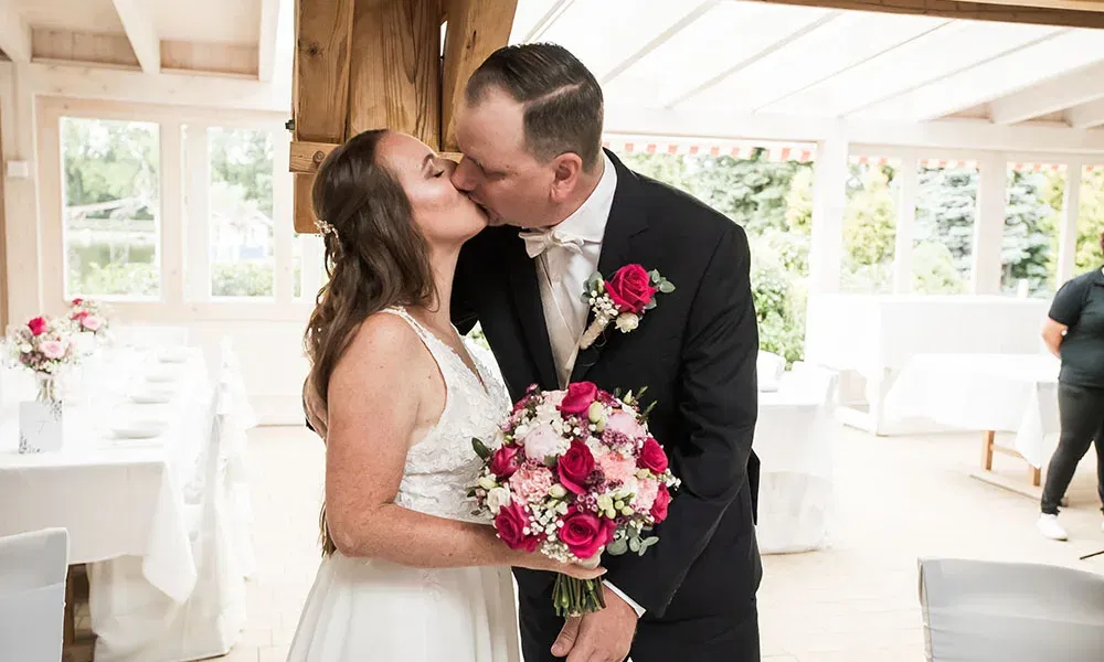 Braut und Bräutigam küssen sich in einer hellen Hochzeitslocation, während die Braut ihren rosa-roten Blumenstrauß hält.