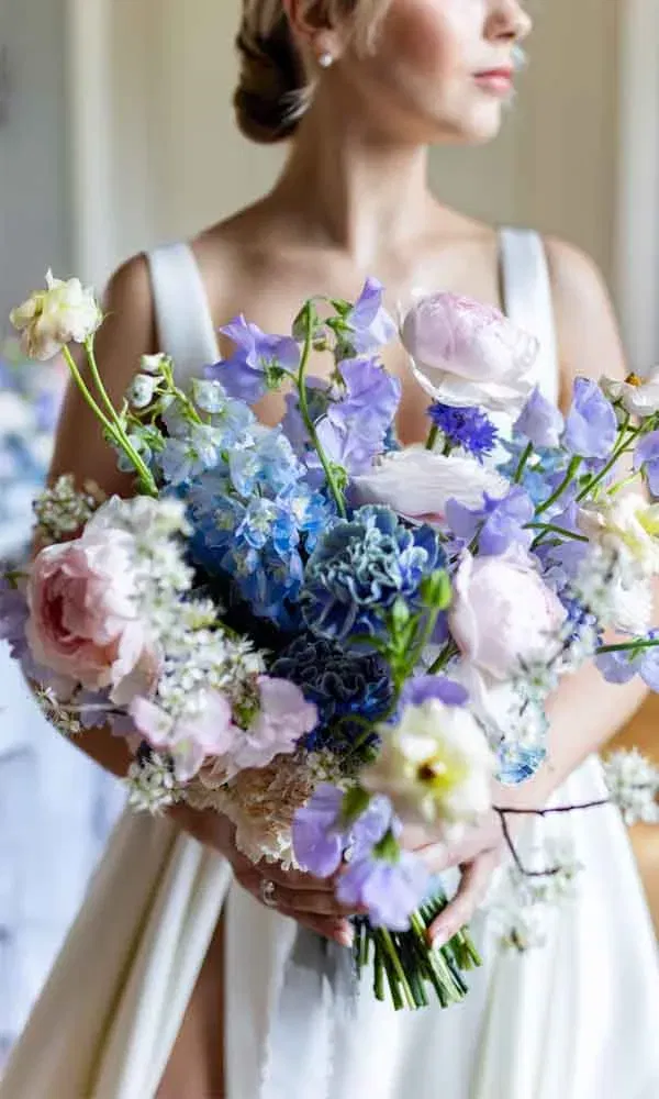 Braut hält einen farbenfrohen Brautstrauß in Blau-, Flieder- und Rosétönen mit Ranunkeln, Hortensien und zarten Frühlingsblumen.