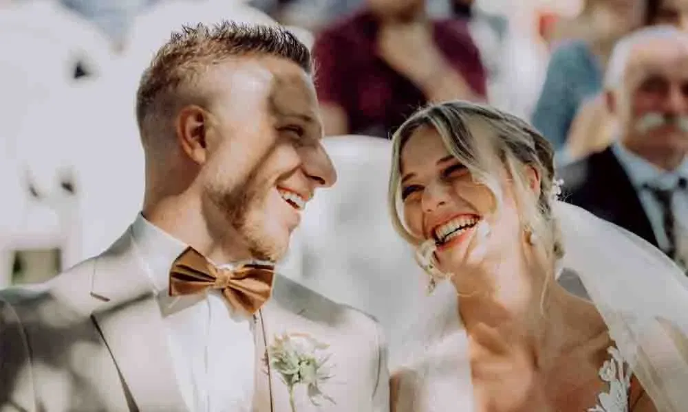 Braut und Bräutigam sitzen nebeneinander und lachen herzhaft während der Trauung, warmes Sonnenlicht fällt auf ihre Gesichter.