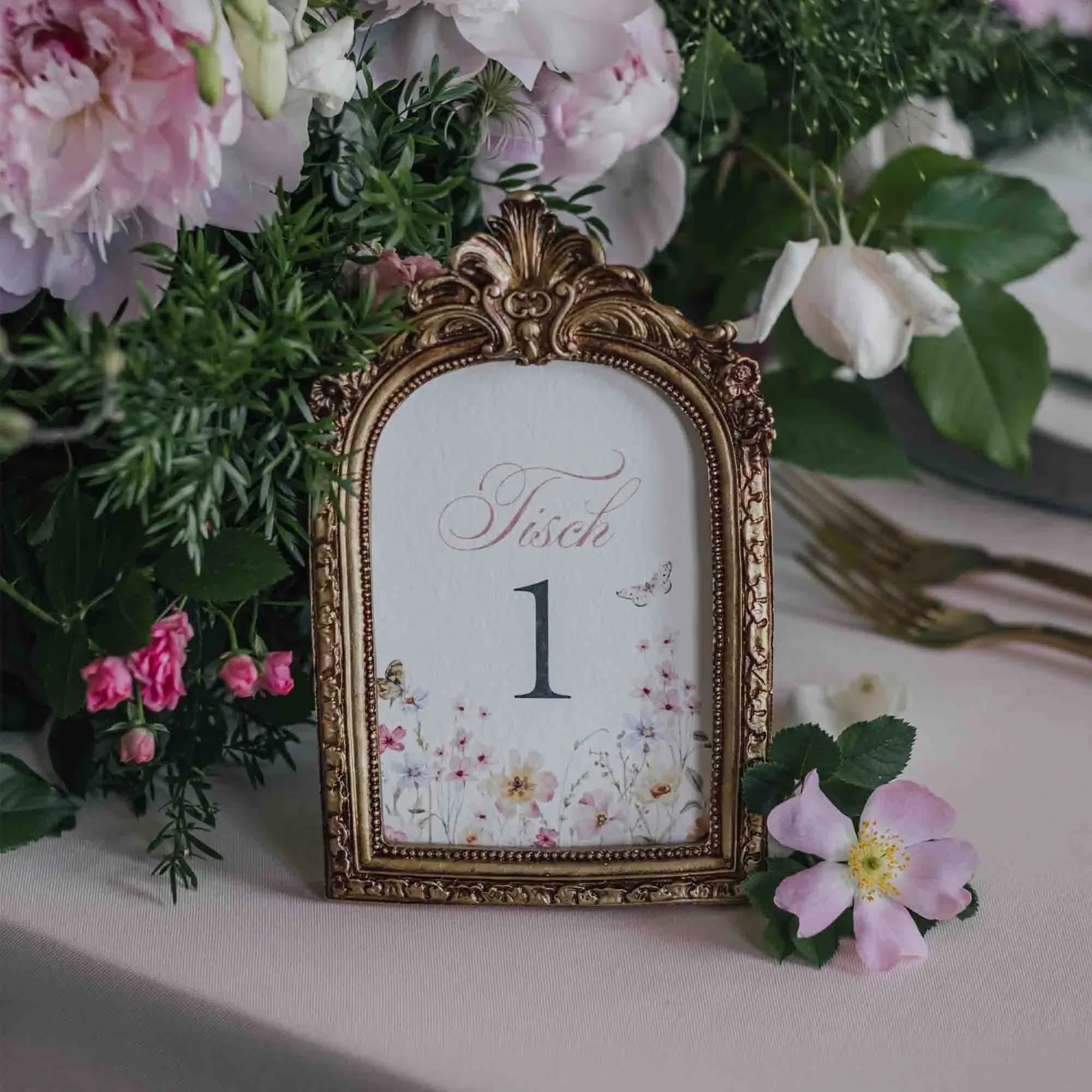 Eleganter Tischaufsteller in goldverziertem Rahmen mit der Aufschrift „Tisch 1“, umgeben von rosa und weißen Blumen in romantischer Hochzeitsdeko.