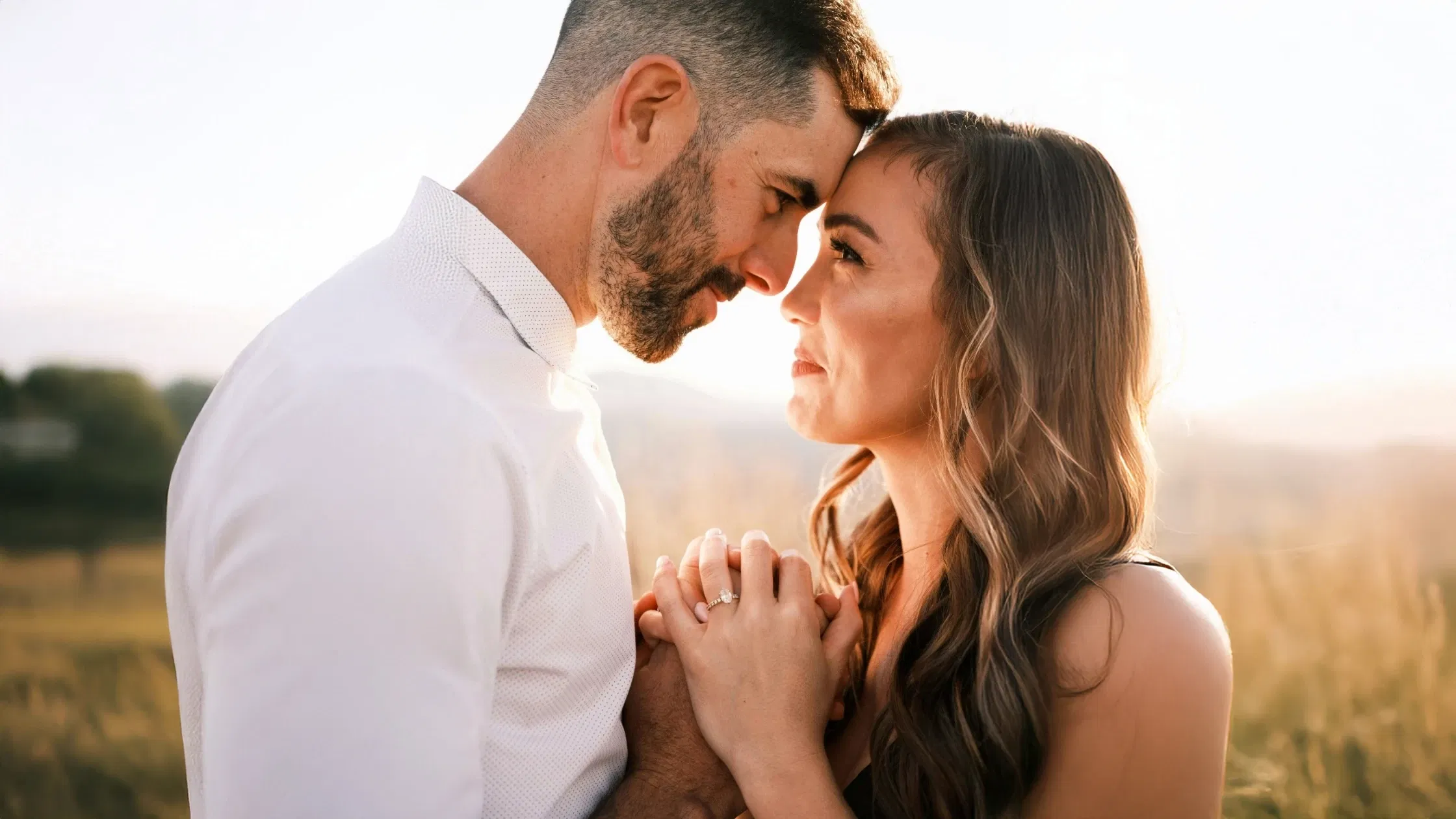 Verlobtes Paar blickt sich verliebt bei Sonnenuntergang auf einer Wiese an.