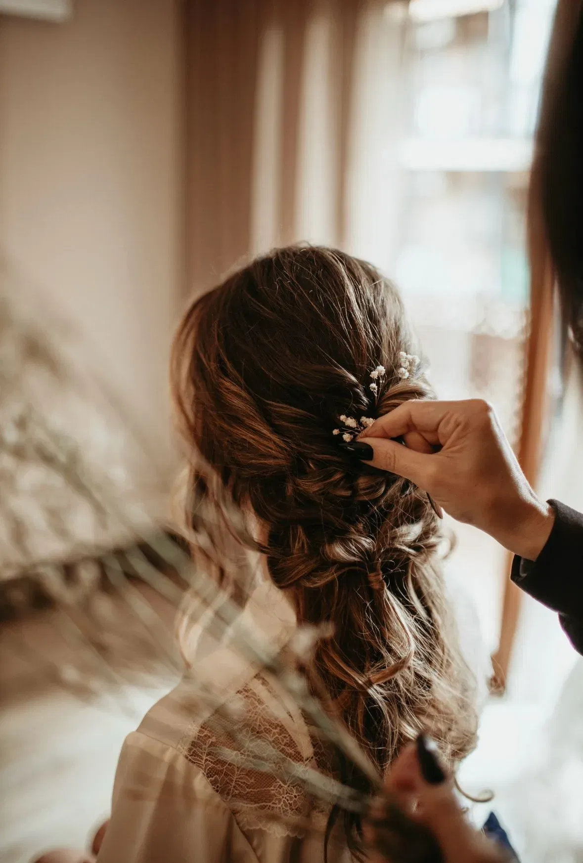 Stylistin steckt kleine weiße Blüten in eine romantisch geflochtene Brautfrisur mit lockeren Wellen, aufgenommen von hinten.