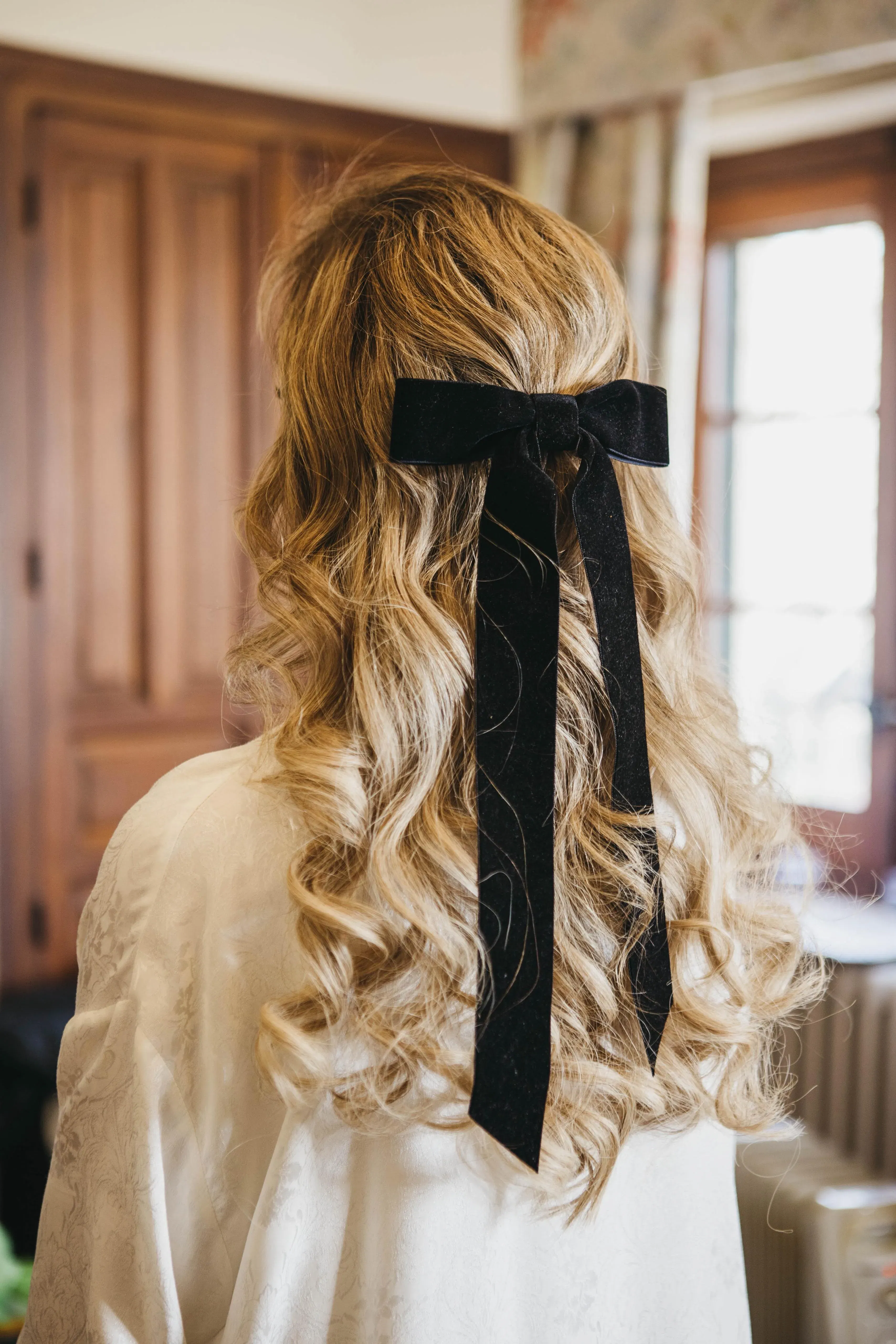 Rückansicht einer Brautfrisur mit weichen, blonden Locken, die mit einer langen schwarzen Schleife im Haar zusammengefasst sind.