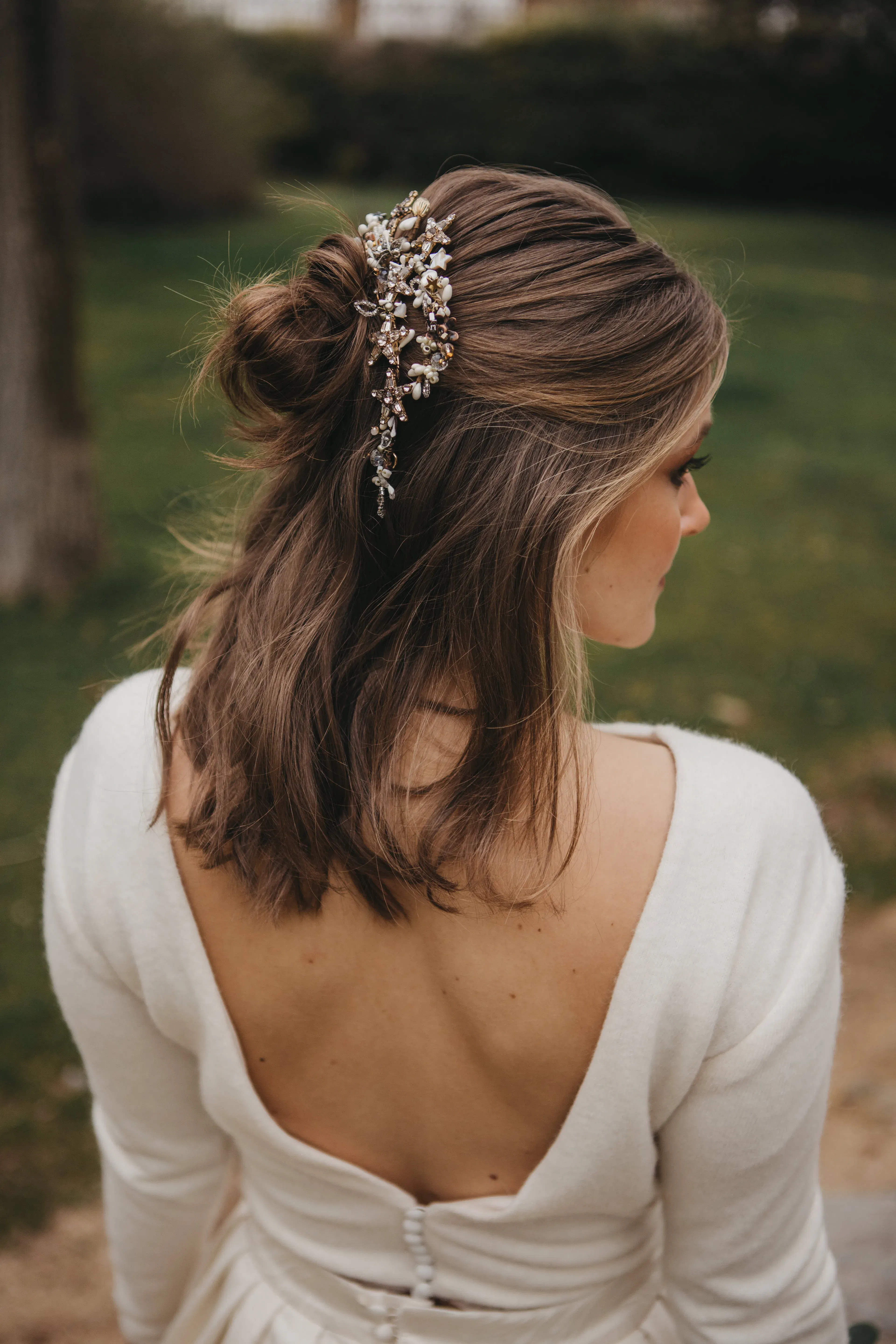 Braut von hinten mit halb offen gesteckter Frisur und floralem Haaraccessoire im Park.