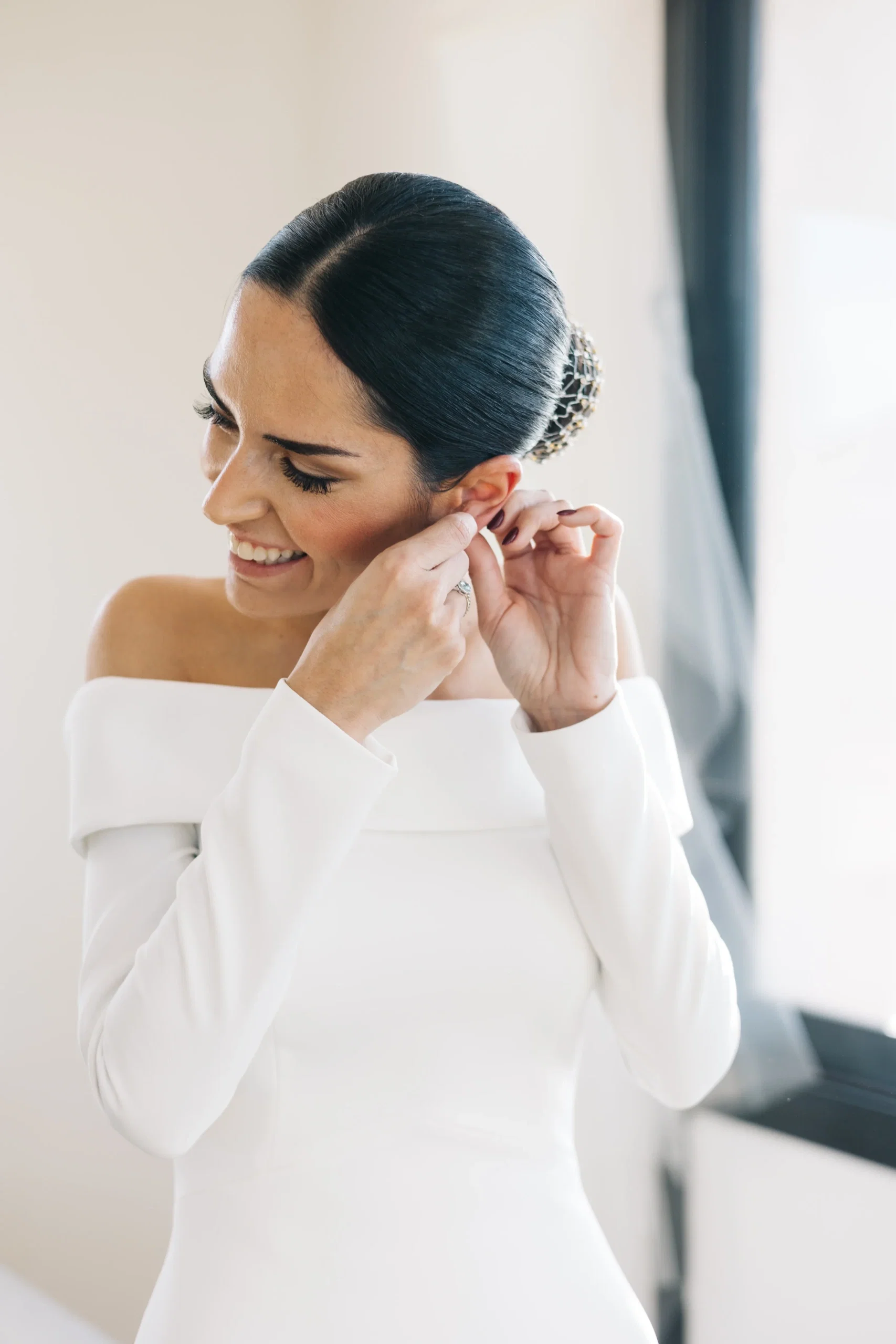 Braut im eleganten Off-Shoulder-Kleid steckt sich einen Ohrring an und lächelt dabei.