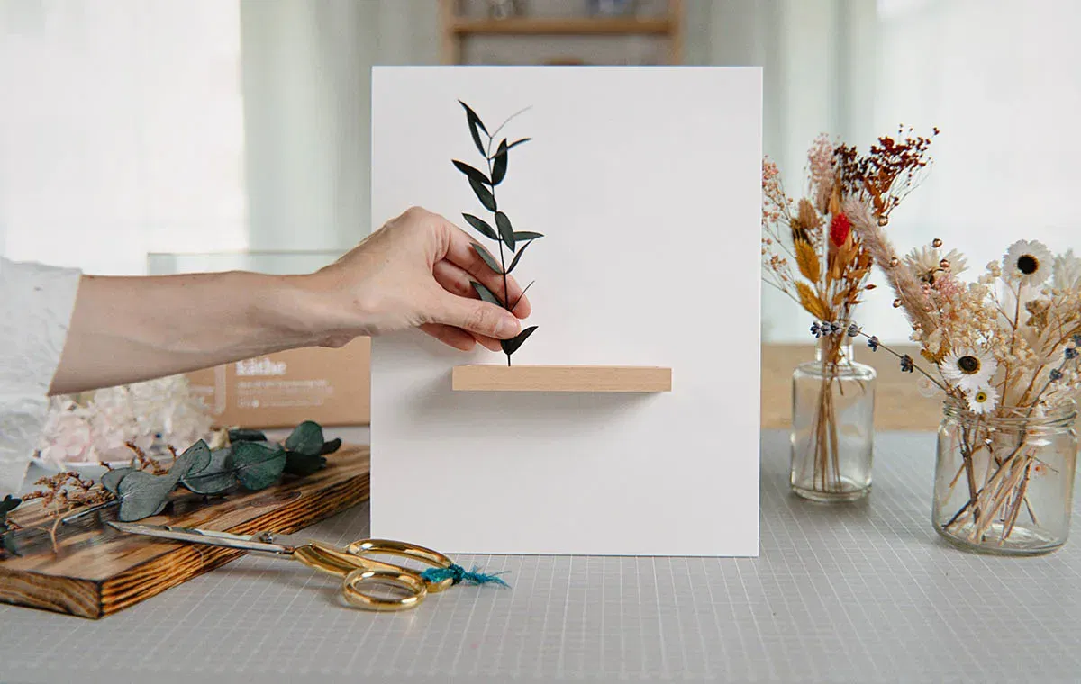 Hand arrangiert einen Eukalyptuszweig auf einem weißen DIY-Bilderrahmen mit kleiner Holzleiste, umgeben von getrockneten Blumen, Schere und Bastelmaterialien.