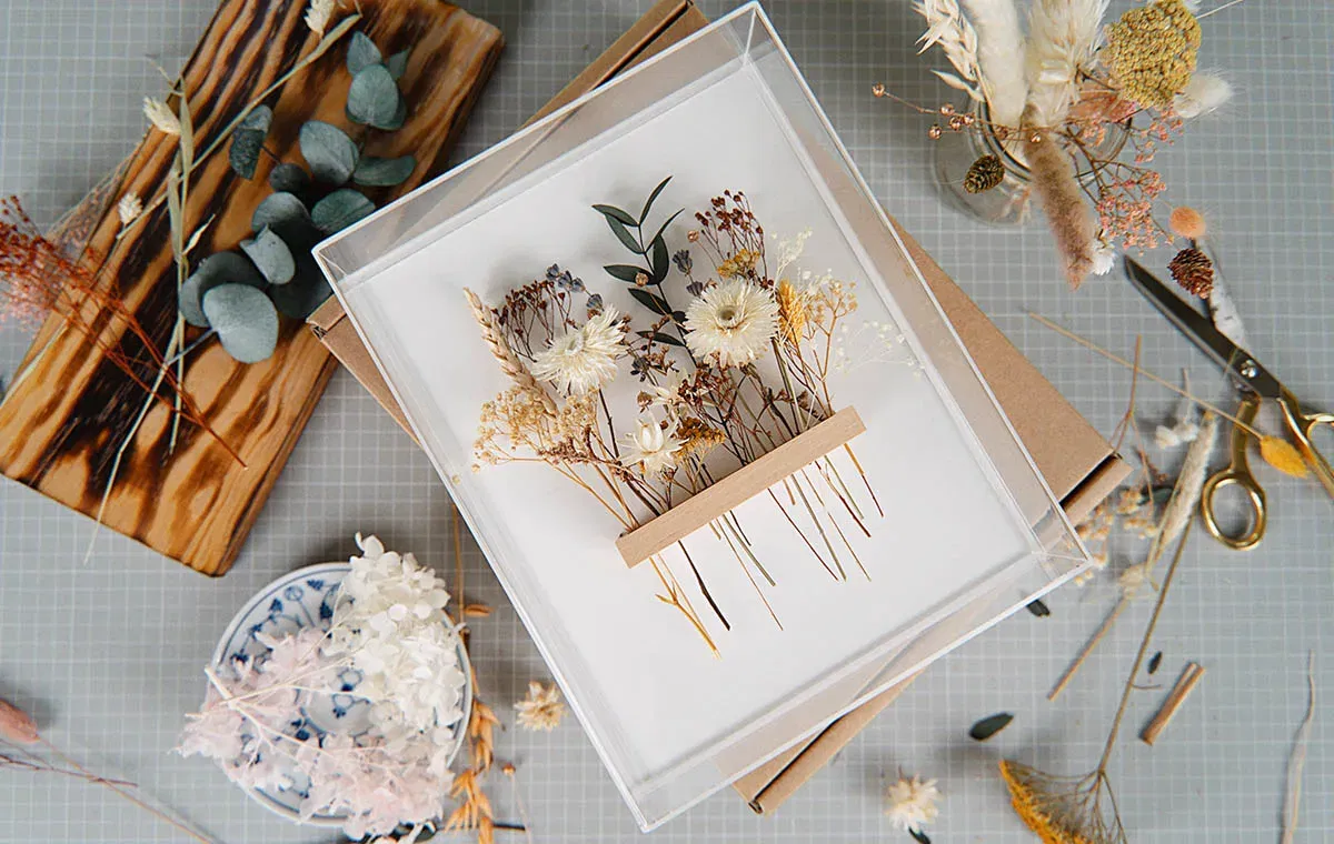 Dekorativer DIY-Bilderrahmen mit zarten getrockneten Blumen in Naturtönen auf weißem Hintergrund, umgeben von Bastelmaterialien und Werkzeugen.