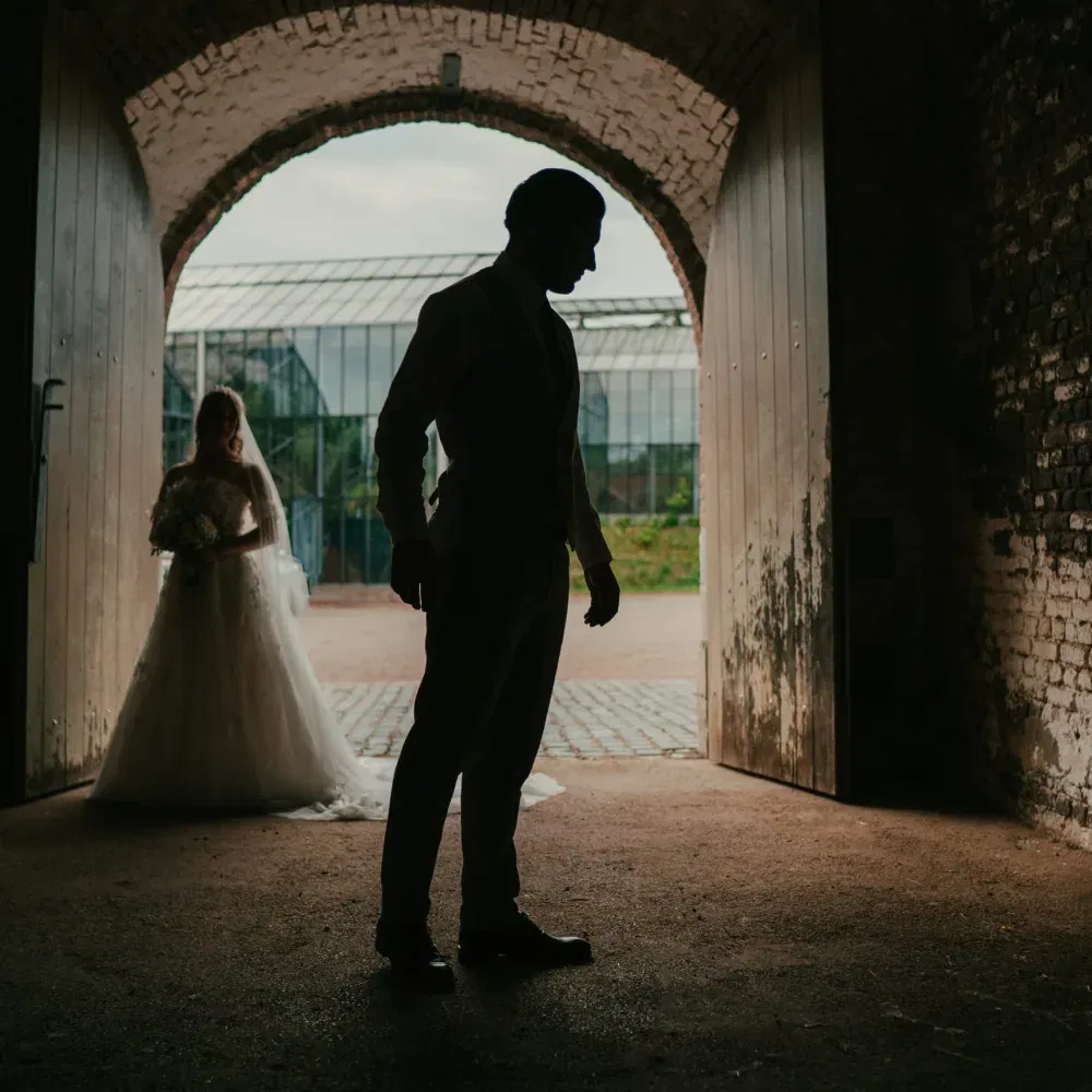 Silhouettenaufnahme eines Bräutigams, der in einem Torbogen steht, während die Braut mit Blumenstrauß im Hintergrund auf ihn zukommt.