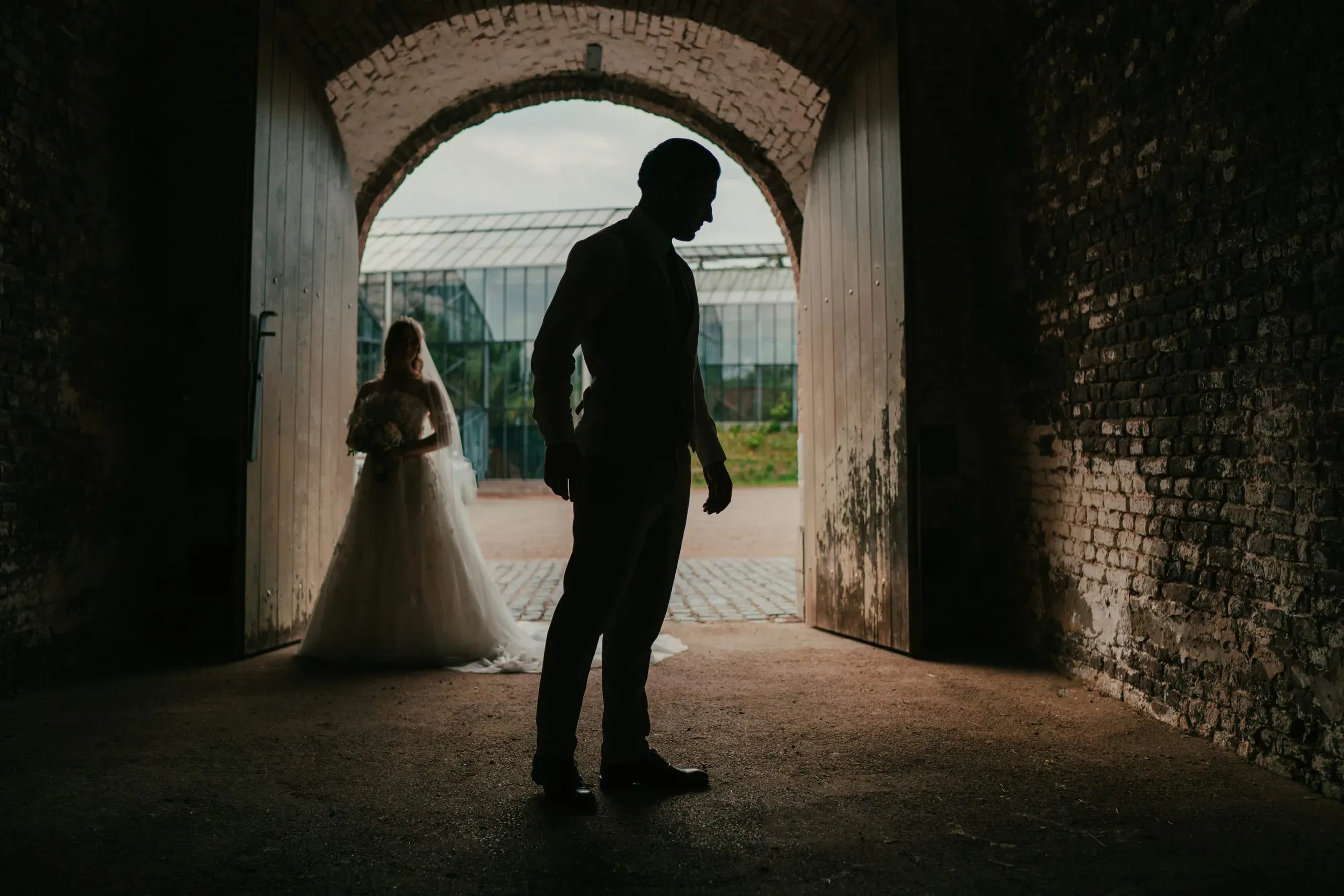 Silhouettenaufnahme eines Bräutigams, der in einem Torbogen steht, während die Braut mit Blumenstrauß im Hintergrund auf ihn zukommt.