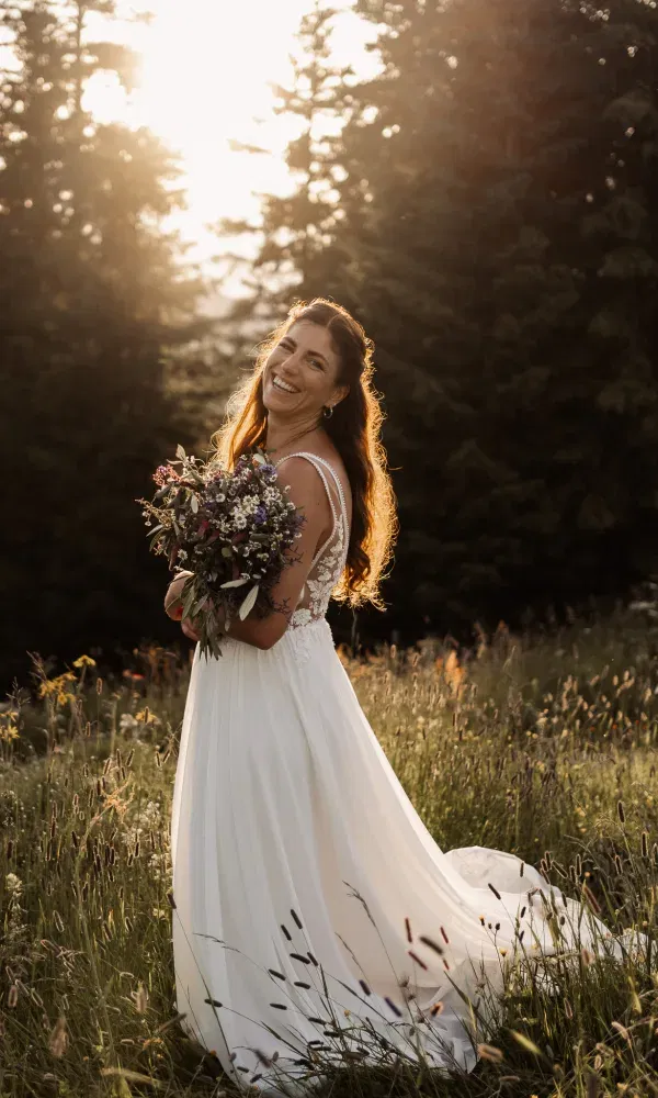 Strahlende Braut im Sonnenuntergang steht auf einer Blumenwiese und hält ihren Brautstrauß.